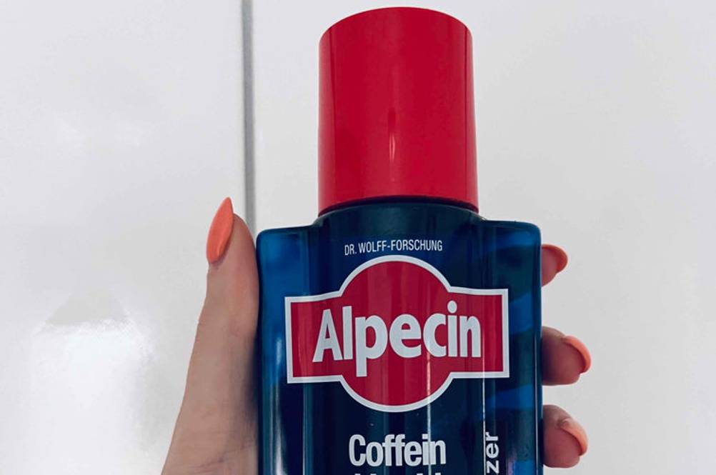Eine Hand hält eine getestete Flasche Alpecin vor eine helle Fließenwand.