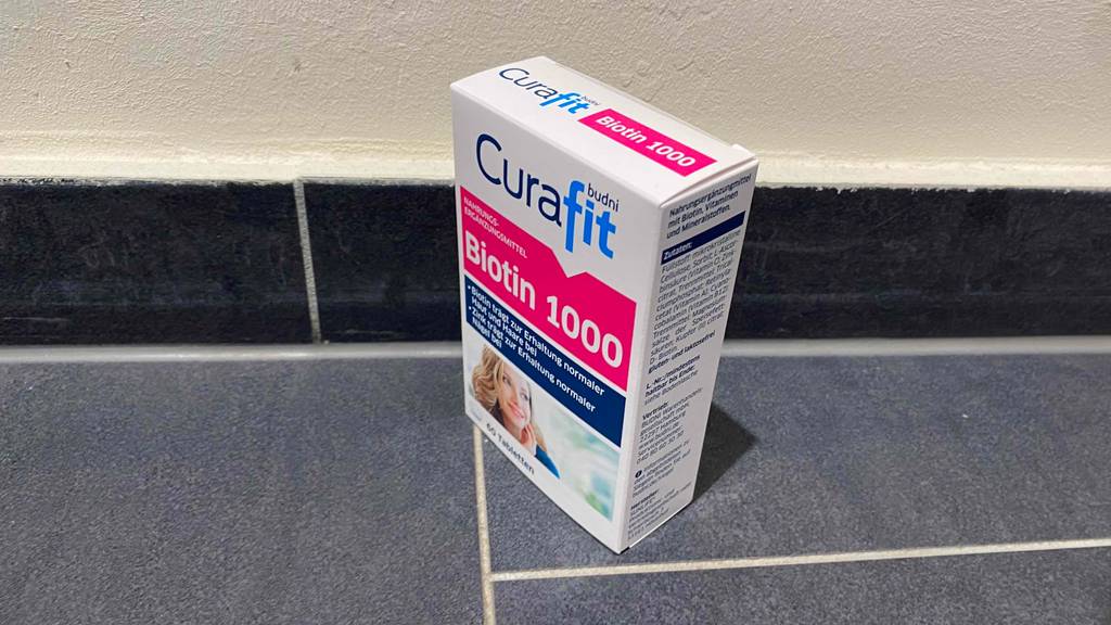 Vor einer hellen Wand steht eine Packung getestetes Biotin auf einem grauen Boden