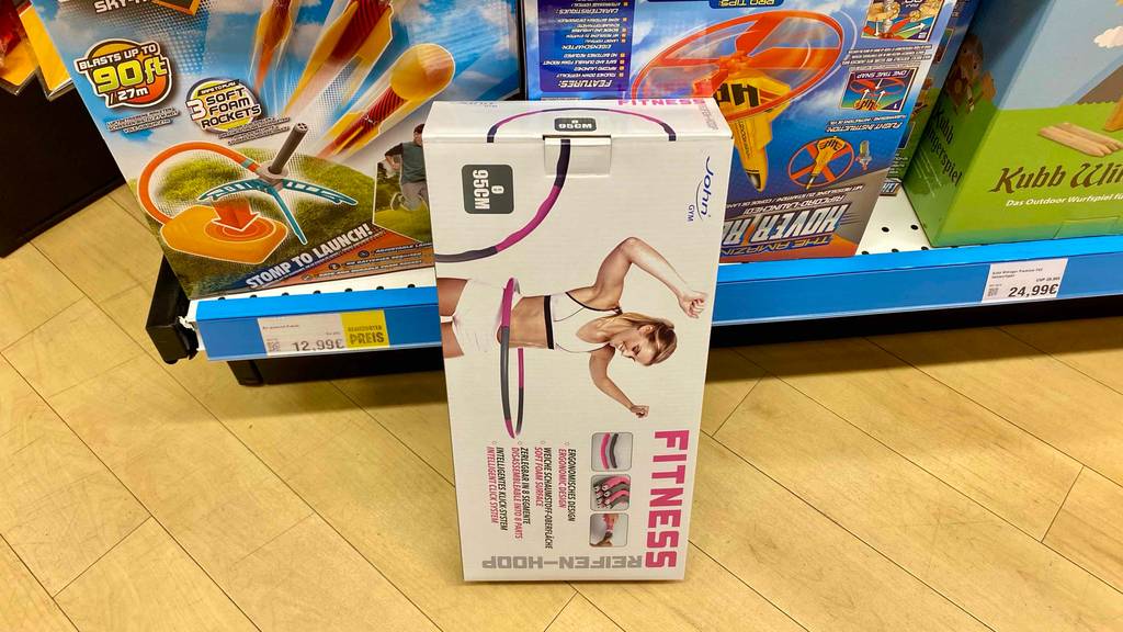 Ein getesteter Hula Hoop Reifen in Originalverpackung lehnt aufgestellt an einem Regal.