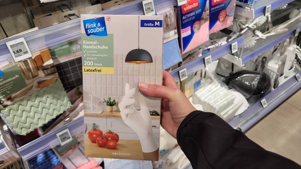 Eine Packung mit getesteten Einmalhandschuhen wird in einer Hand vor einem Regal gehalten.