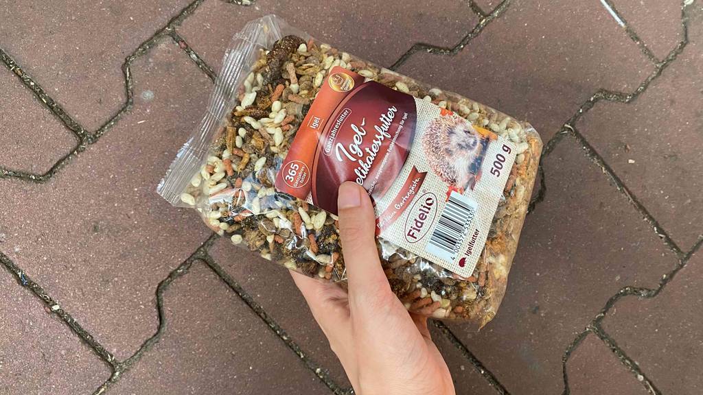 Hand hält getestetes Igelfutter in transparenter 500-g-Packung von Fidelio inklusive Aufkleber mit Beschreibung über rötlichem Steinbelag.