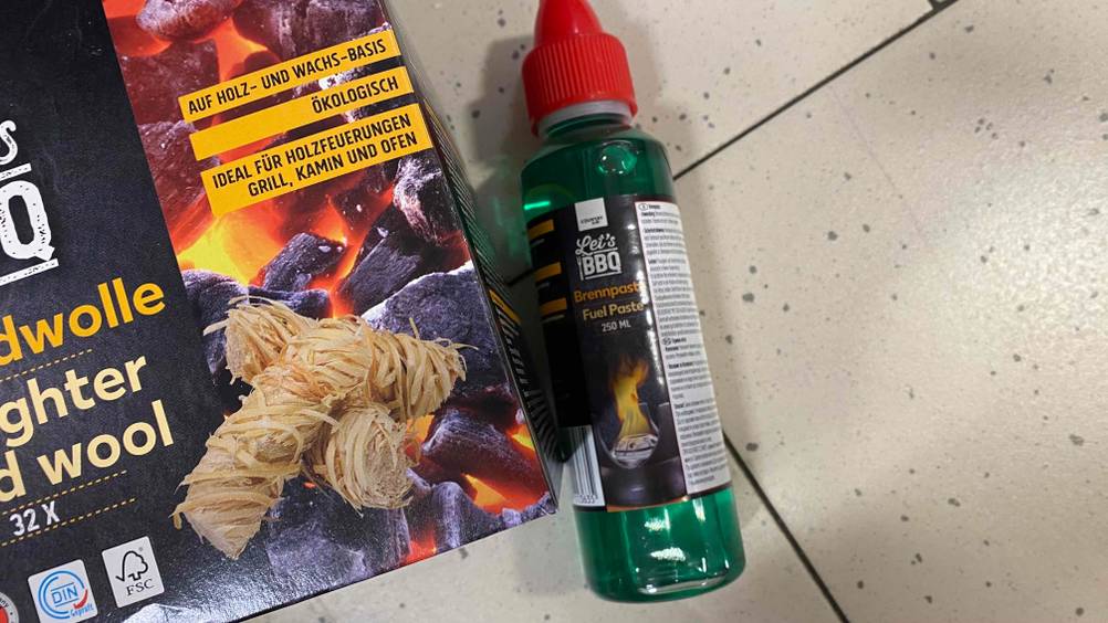 Eine Flasche Brennpaste neben einer Packung Grillwolle auf einem Fließenboden.