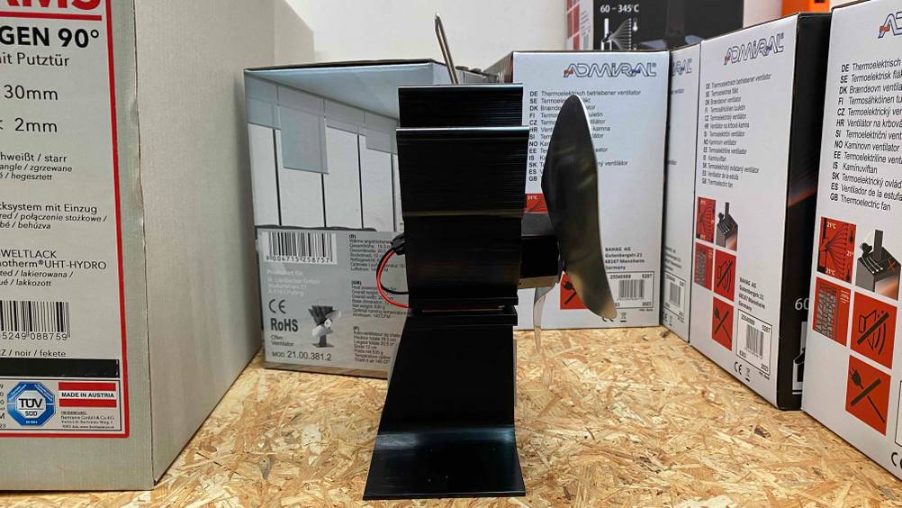 Ein getesteter Ofenventilator von der Seite auf einem Holzregal neben verpackten Geräten.