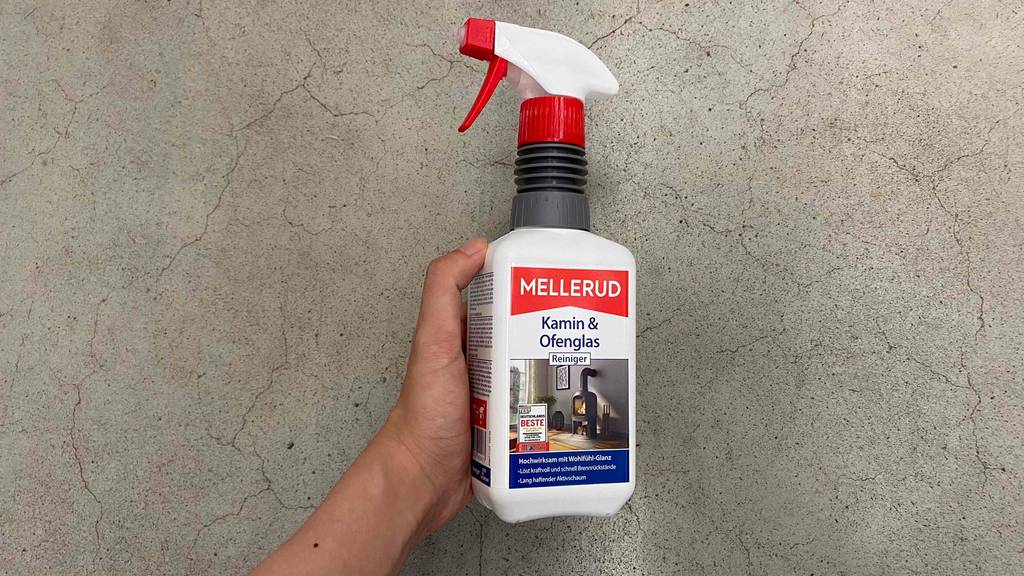 Eine Hand hält eine 500-ml-Sprühflasche inklusive getestetem Mellerud-Kaminreiniger vor hellgrauem Hintergrund.