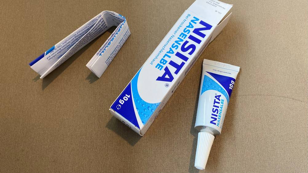 Im Test: Alle Bestandteile einer Packung Nasensalbe, Tube, Beipackzettel und Verpackung auf eiener beigen Fläche.