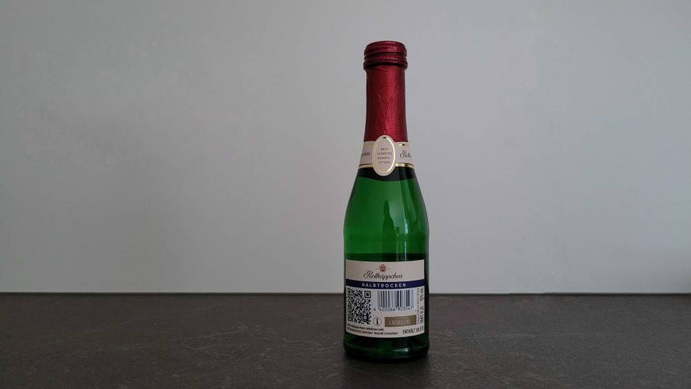 Sekt im Test: Eine kleine Flasche Sekt steht vor einer hellen Wand, Rückseite nach vorne