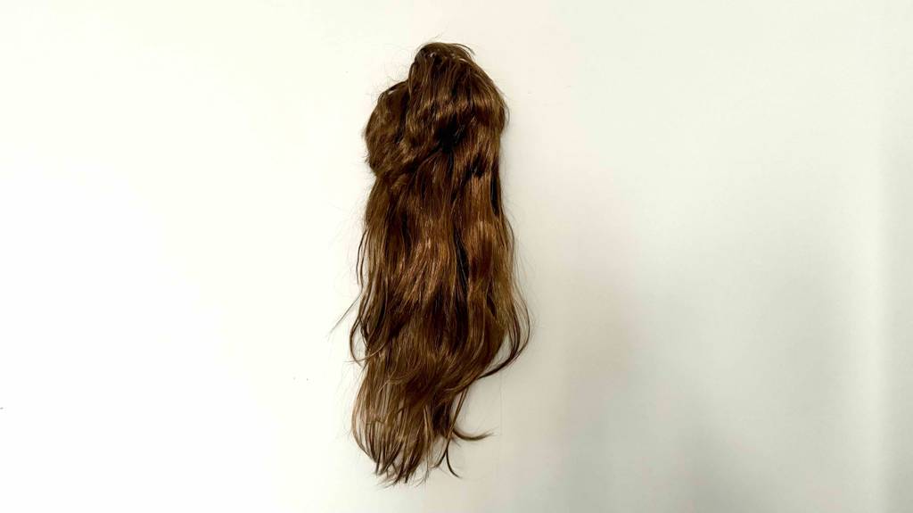 Test: eine braune Perücke mit langen Haaren vor einer weißen Wand im Fokus.