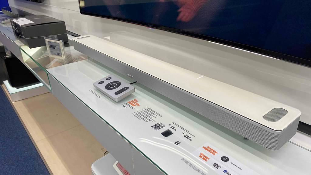 Eine getestete Soundbar (weiß) von der Seite unter einem Fernseher auf einer hellen Fläche.