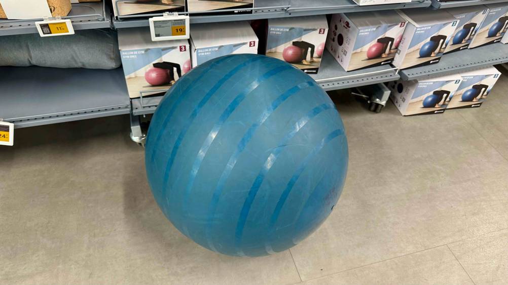 Der getestete 65 cm Gymnastikball in der Farbe Blau ist vor einem Regal platziert.