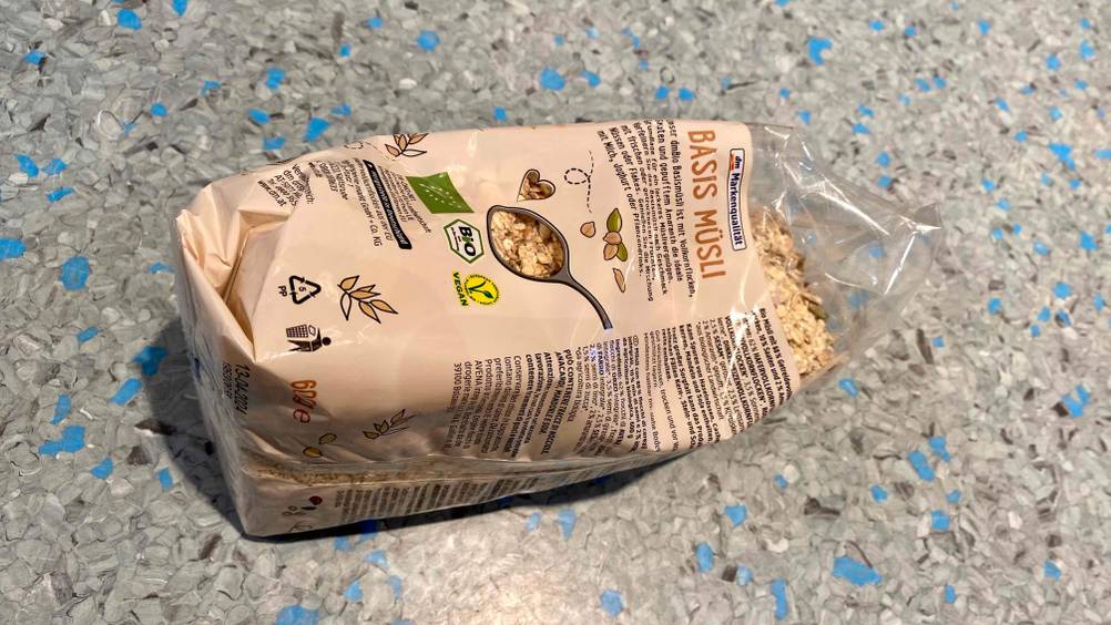Eine Packung getestetes Basis-Müsli von schräg hinten auf einer grauen Fläche liegend.
