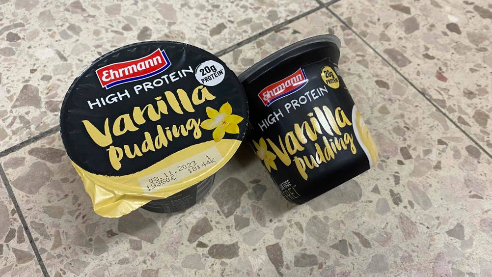 Protein Pudding im Test: Zwei Becher Pudding einer liegend einer stehend auf einer Fließenoberfläche