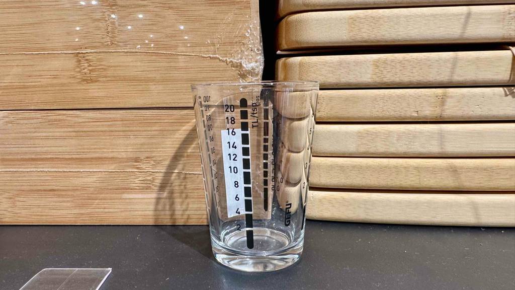 Ein Messbecher mit schwarzer Skala, der getestet wurde steht vor Holzgegenständen.