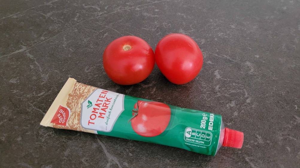Eine getestete Tube Tomatenmark neben zwei Tomaten auf einer dunklen Fläche.