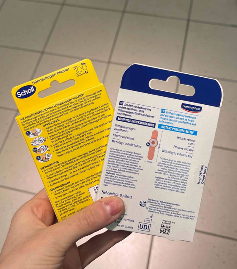 Hühneraugenpflaster-Test: Zwei verschiedene Verpackungen werden mit der Rückseite ersichtlich in der Hand gehalten.