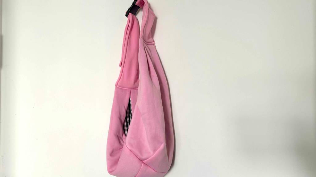 Hundetragetuch im Test: ein rosa Hundetragetuch mit verschlossener Schnalle hängt vor einer weißen Wand.