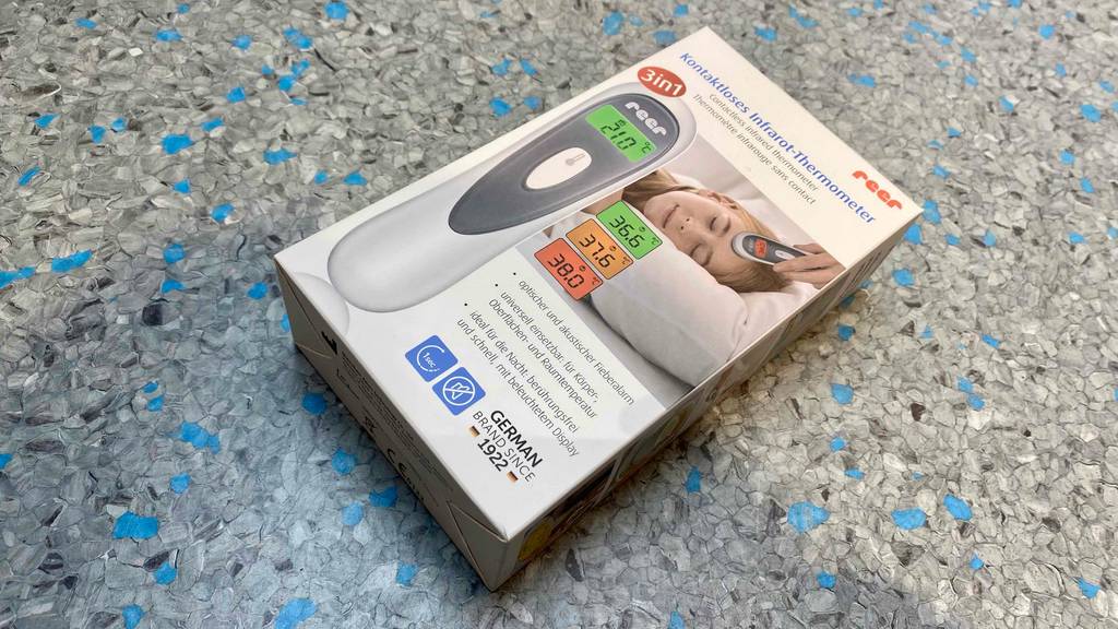 Auf einem grauen Boden liegt quer ein getestetes Reer-Fieberthermometer in Originalverpackung.