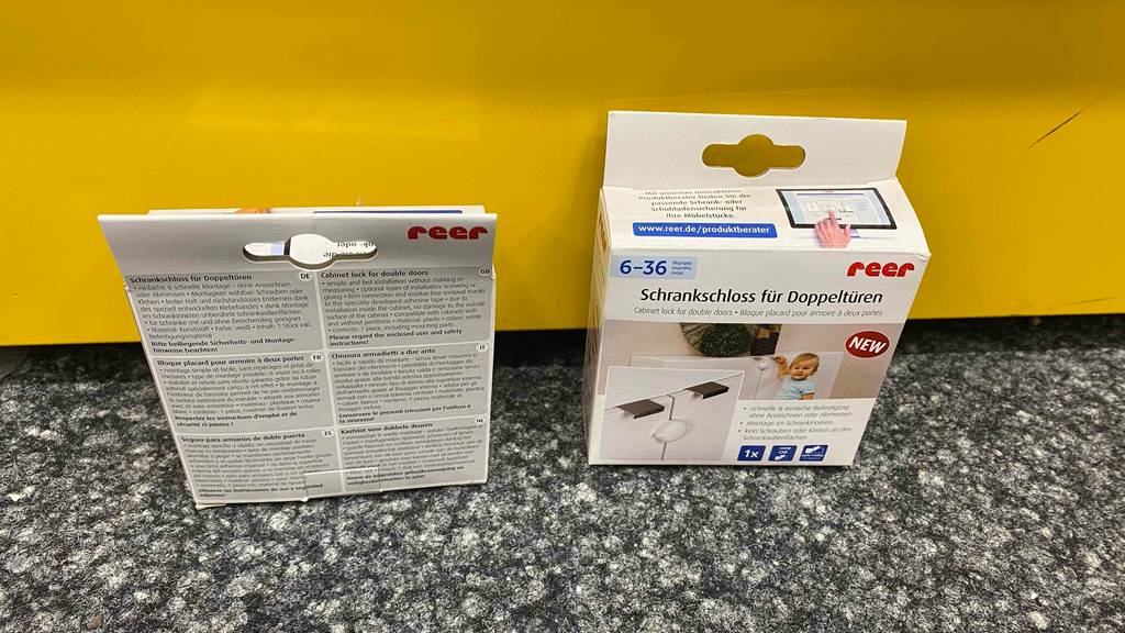 Zwei Packungen der getesteten Schrank-Kindersicherung vor einer gelben Wand auf einem grauen Teppich.