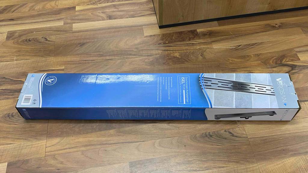 Auf einem Boden liegt eine Originalverpackung einer getesteten Duschrinne horizontal auf einem Holzboden.