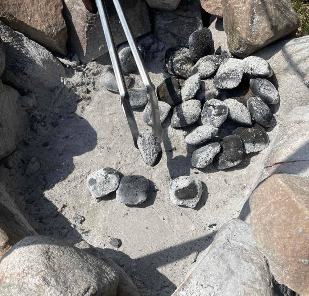 Grillbriketts im Test: Grillbriketts sind auf einer Feuerestelle platziert, währenddessen eine in einer Zange gehalten wird.