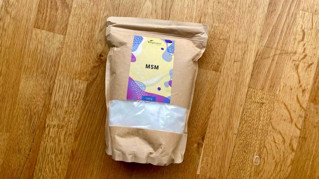 MSM-Pulver im Test: Das Pulver in einer braunen Papiertüte mit Sichtfenster liegt auf einer Holzfläche.