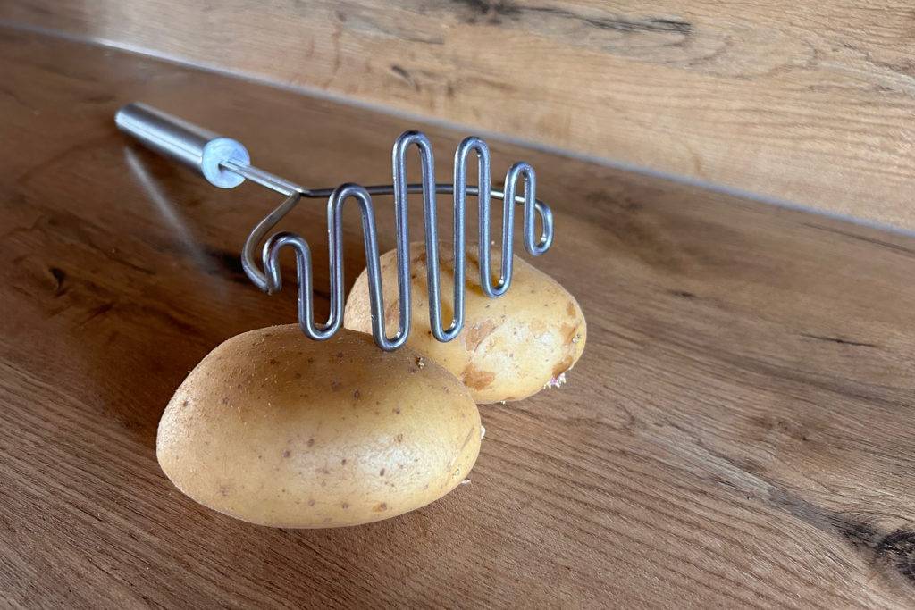 Kartoffelstampfer und zwei Kartoffelhälften auf einer Holzoberfläche.