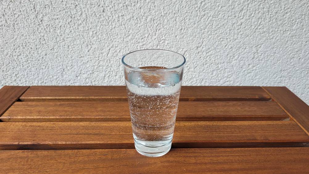 Im Test: Ein Glas Adelholzener-Wasser steht auf einer Holzoberfläche vor einer hellen Wand.