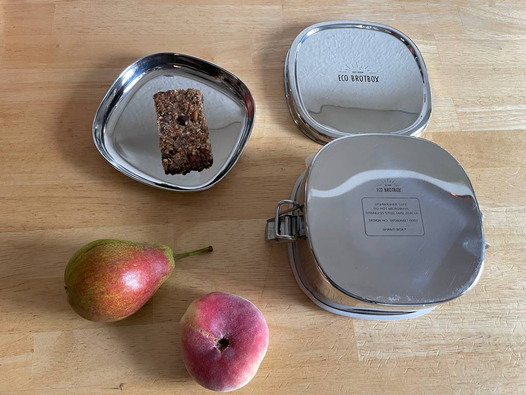 Die geöffnete und getestete Brotdose aus Metall liegt mit Lebensmitteln auf einem Holztisch.