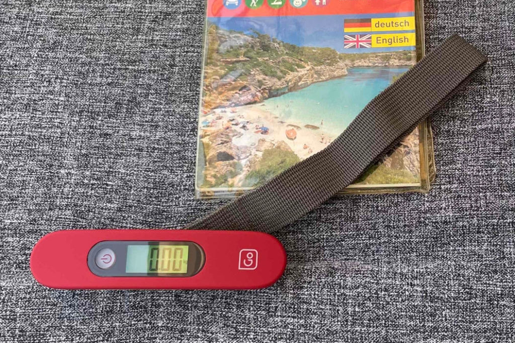 Getestete Kofferwaage mit beleuchteter Gewichtsanzeige und Haltegurt sowie Mallorca-Freizeitkarte auf graumeliertem Untergrund.