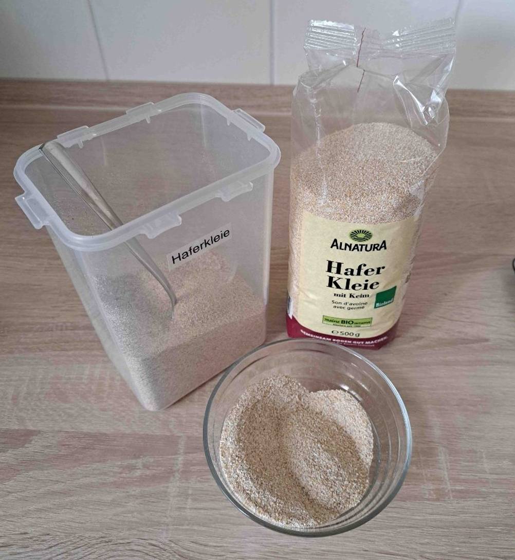 Haferkleie im Test: eine Schüssel mit Haferkleie darin, steht neben der Verpackung und einem Aufbewahrungsbehältnis, worin sich ebenfalls Haferkleie befinden.
