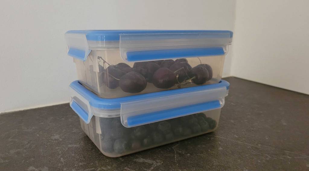 Zwei verschlossene Frischhaltedosen gestapelt mit Kirschen und Heidelbeeren darin.