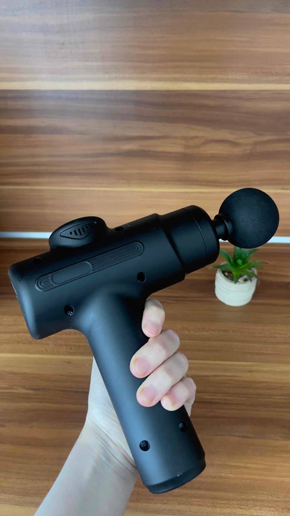Eine Hand hält die getestete Mini-Massagepistole über einem Kaktus im Topf.