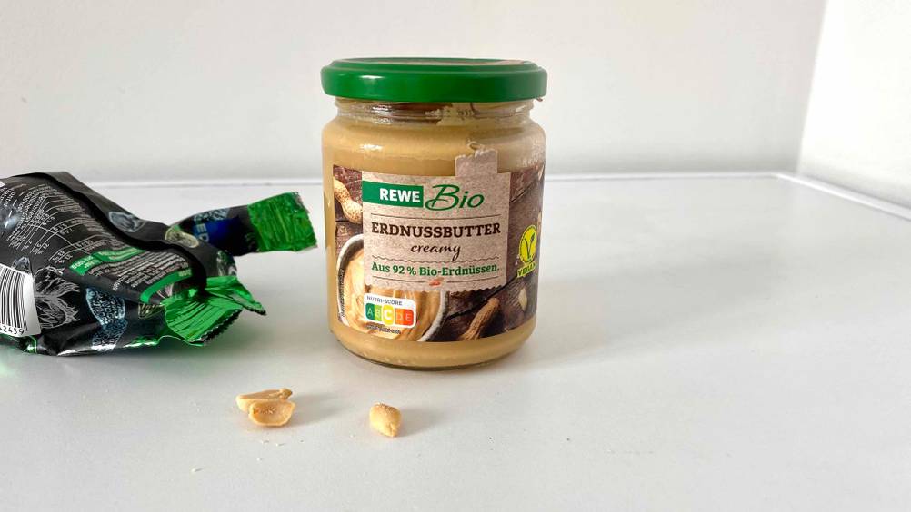 Ein Glas getestete Erdnussbutter neben einer Packung Erdnüssen auf einer hellen Fläche.