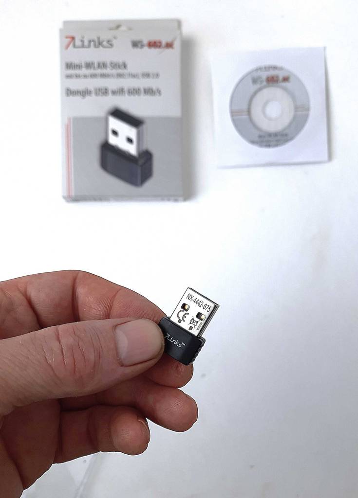 Getestet: Der WLAN-Stick 5 GHz wird von einer Hand gehalten, im Hintergrund befinden sich die Installations-CD inklusive der WLAN-Stick-Verpackung.