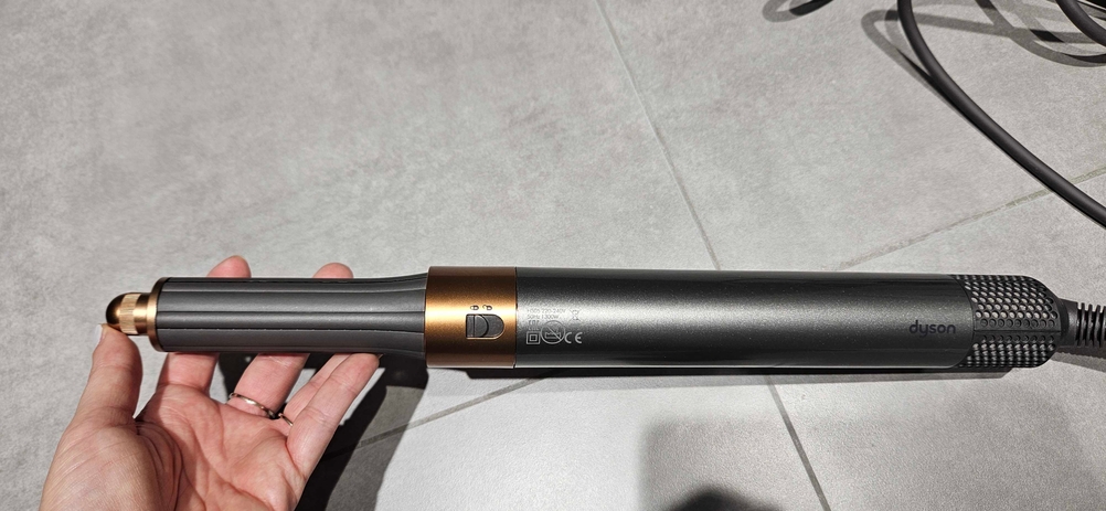 Eine Hand hält einen getesteten Lockenstab vor eine graue Fließenfläche.