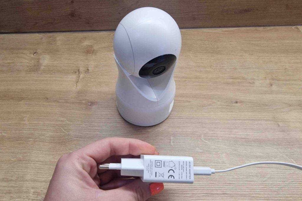 Weißes Babyphone mit Kamera stehend und davor wird ein Ladekabel in der Hand gehalten.