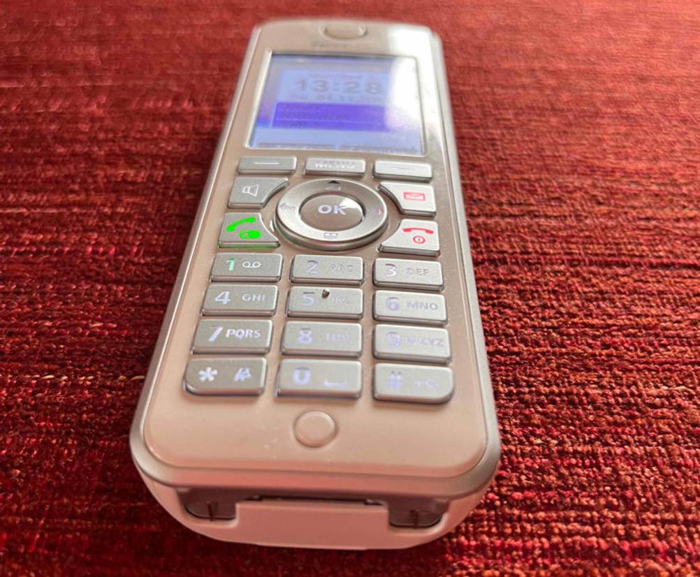 Weißes Fritz-IP-Telefon im Test liegt eingeschaltet mit Fokus auf die Aufladevorrichtung im unteren Bereich auf rotem Untergrund