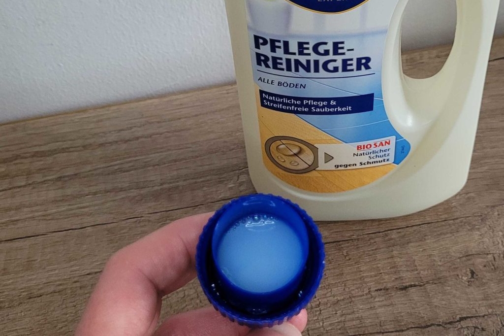 Bodenreiniger in Dosierkappe steht auf dem Boden. Eine Hand hält die Dosierkappe,