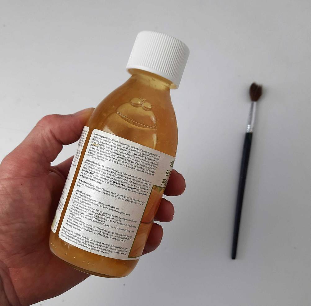 Das getestete Holzöl wird in der Hand gehalten, während auf der Verpackung verschiedene Hinweise und Informationen zum Produkt stehen.