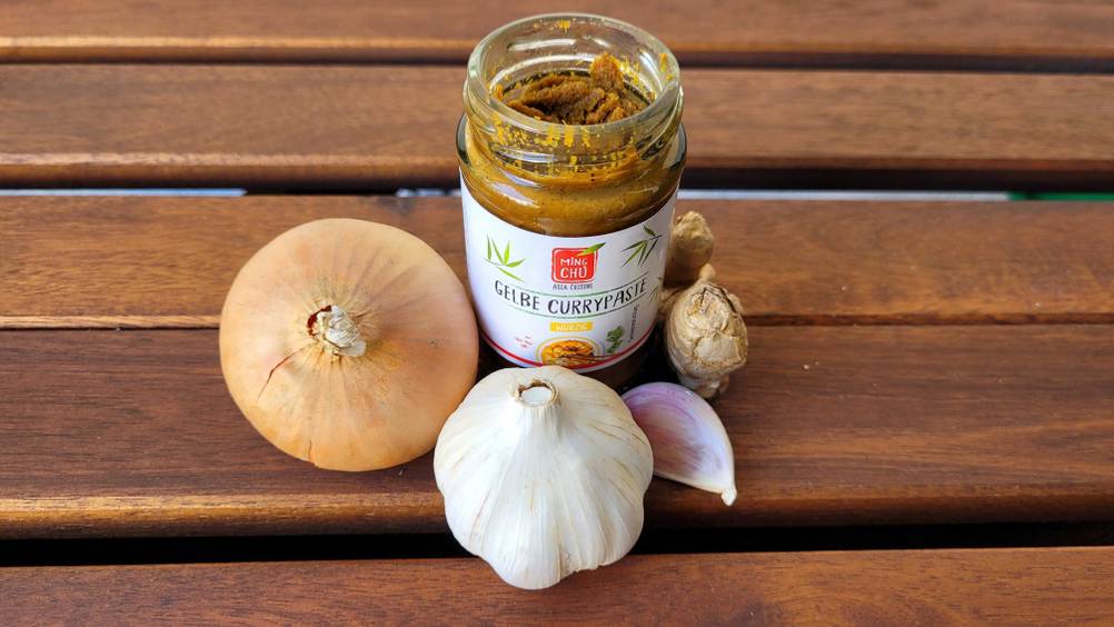 Im Test: Ein Glas gelbe Currypaste steht neben Zwiebel und Knoblauch auf einer Holzfläche.