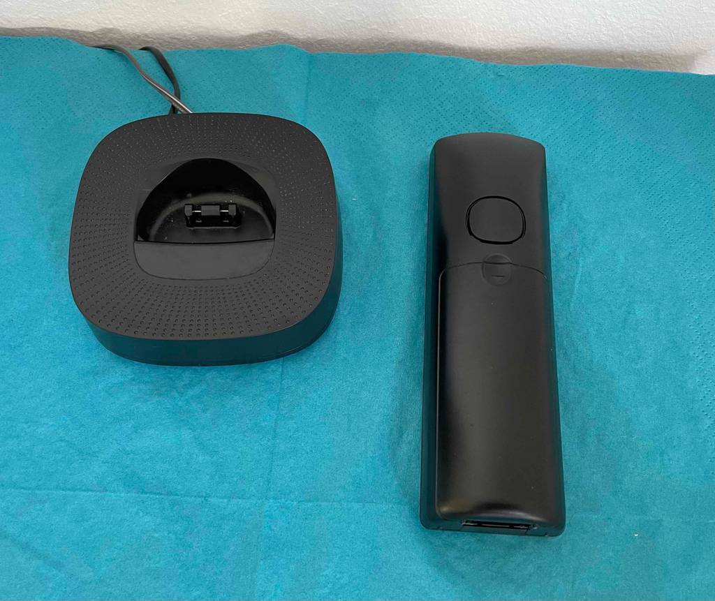 Getestetes schnurloses Telefon liegt mit dem Batteriefach nach oben auf türkisfarbenem Untergrund neben seiner Ladestation.