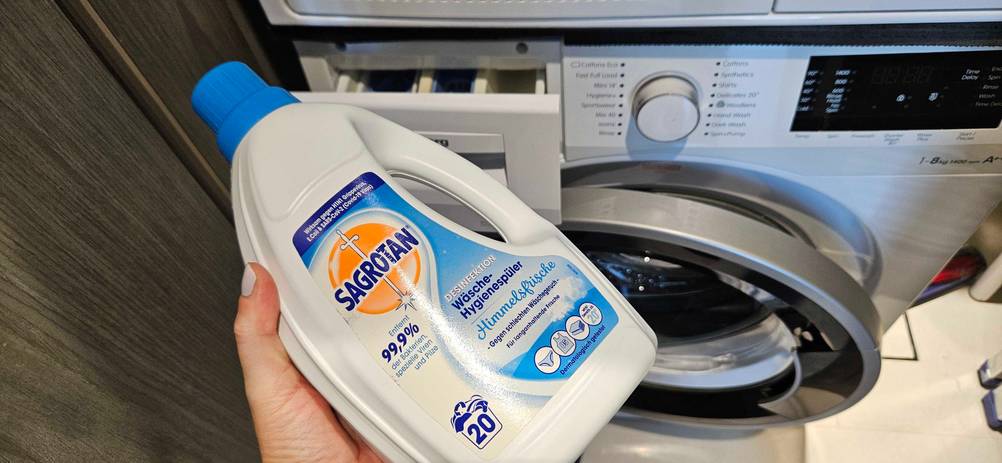 Eine Hand hält eine Flasche Hygienespüler vor eine Waschmaschine.