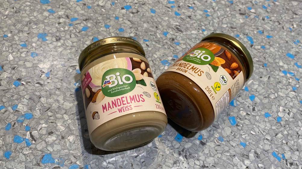 Zwei Gläser getestetes Mandelmus liegen nebeneinander auf einer grauen Fläche.