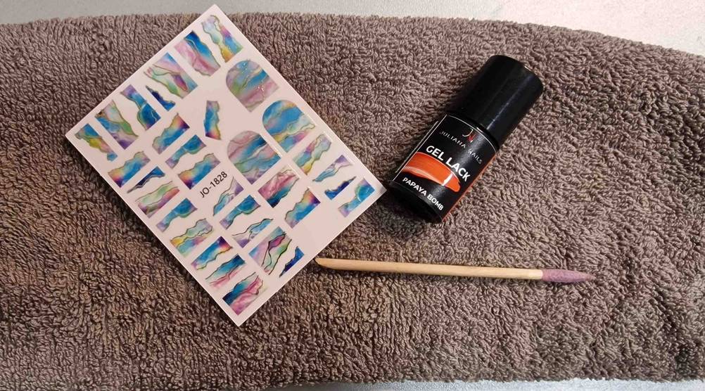 Gel-Nagellack im Test: Ein Gel-Nagellack in der Farbe Orange, Schablonen und ein Nagelhautschieber auf einem Handtuch platziert.