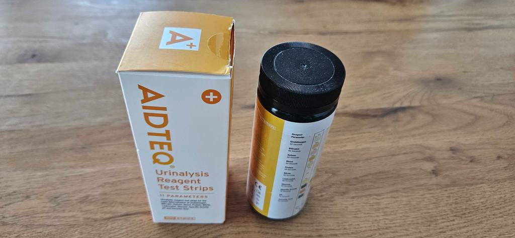 Urinteststreifen getestet: Eine Dose und eine Verpackung nebeneinander auf einer Holzoberfläche stehend.