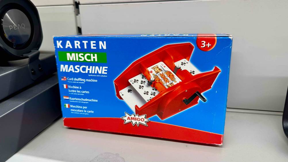Die getestete Kartenmischmaschine mit Kurbel steht in einem Regal neben einem anderen Artikel.