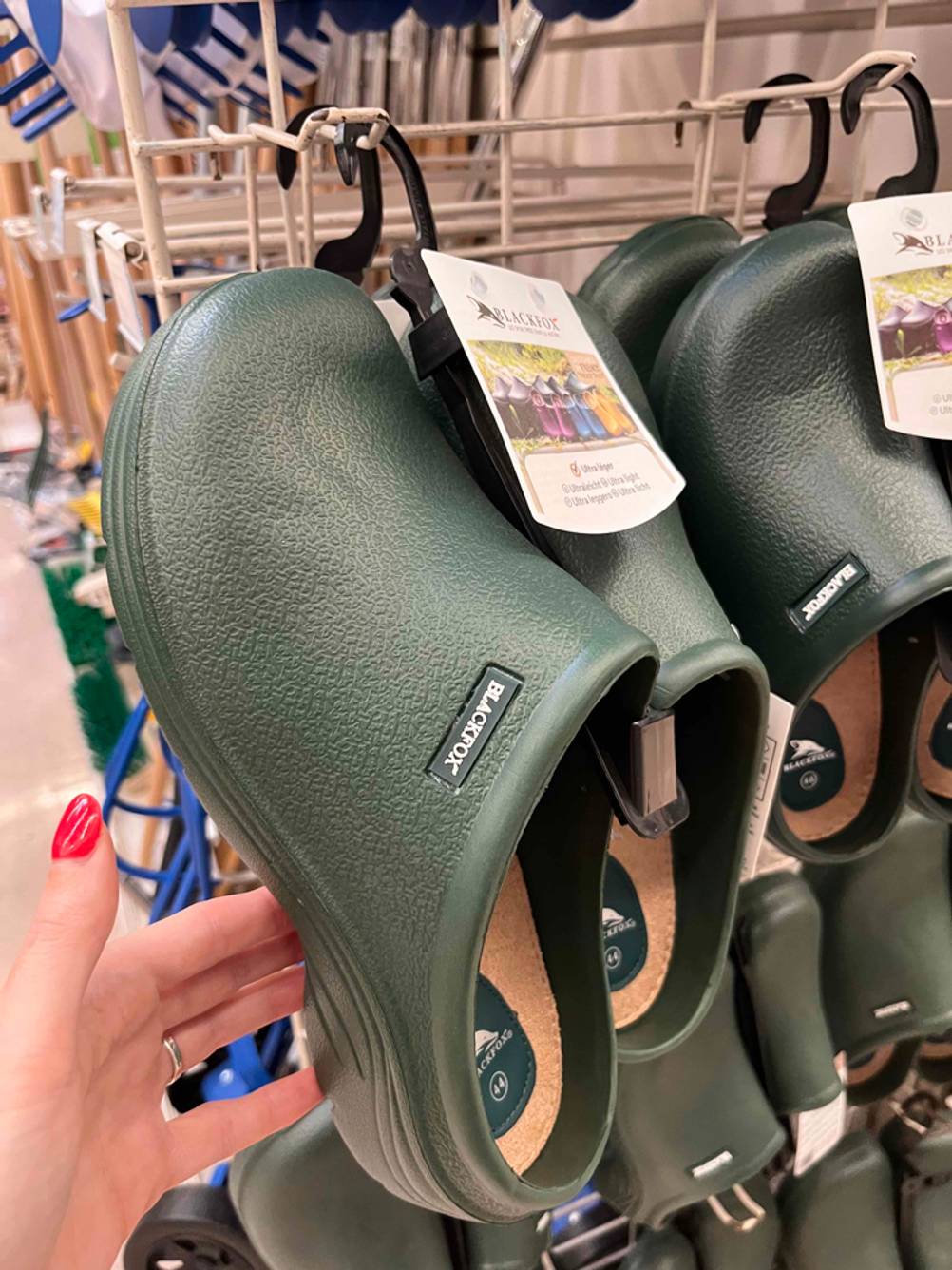 Gartenschuhe im Test: Mehrere Paare hängen an einem Gitter, eine Hand hebt einen Schuh an