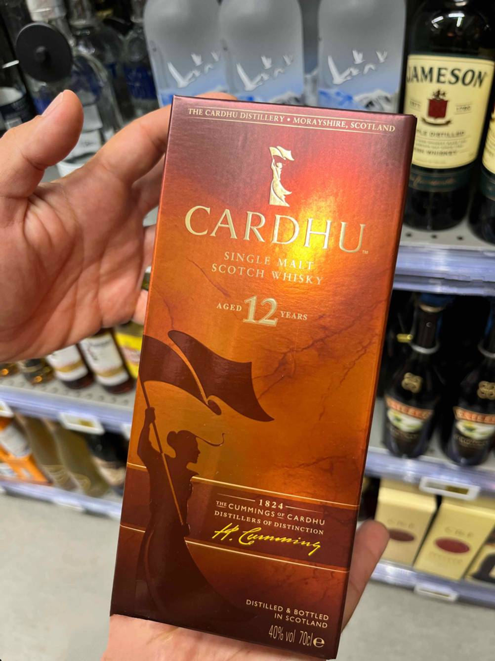 Single-Malt-Scotch-Whisky getestet: Eine Hand hält eine Packung Whisky nach oben im Hintergrund ein Regal