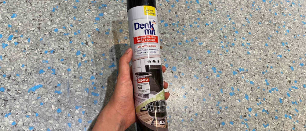 Grillreiniger im Test: Eine Hand hält eine Dose vor Fließenboden