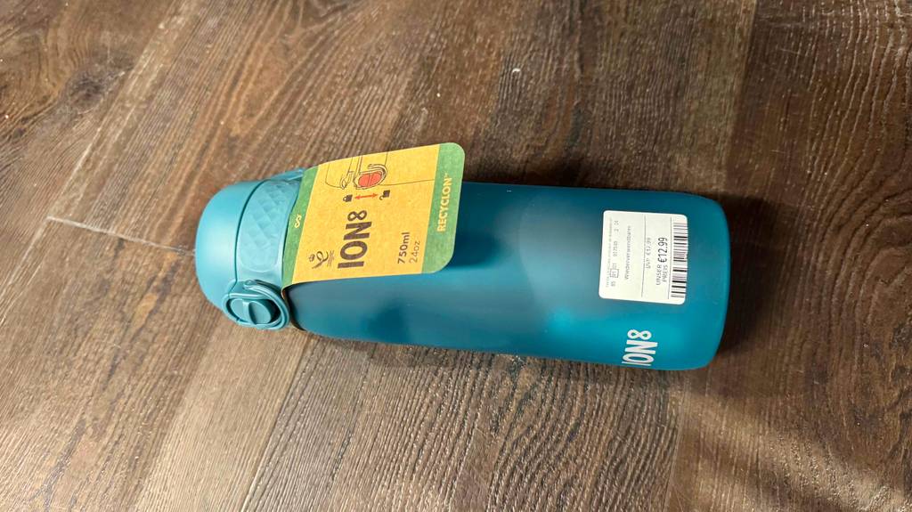 Die getestete Kunststoff-Trinkflasche liegt auf einem Boden an der ein Etikett befestigt ist, mit Infos zum Produkt.