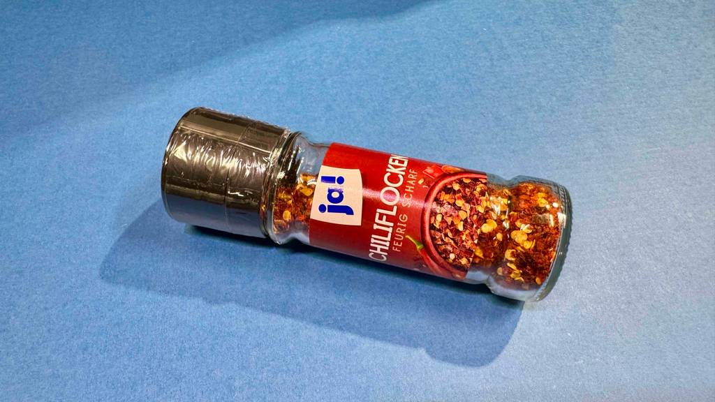 Getestete Chiliflocken liegen im Glas auf einer blauen Fläche.